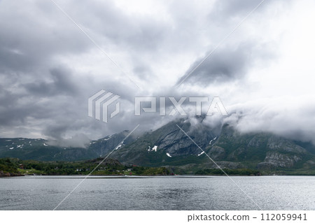 Summer Norwegian fjord where low clouds meet towering mountains and green shores mirror moody skies 112059941