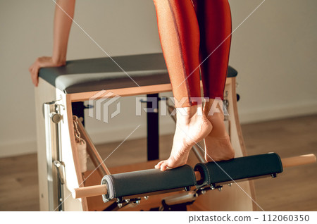 Woman in red leggings does exercises to strengthen legs muscles on Pilates reformer in sports studio Woman in red leggings does exercises to strengthen legs muscles on Pilates reformer in sports studio 112060350