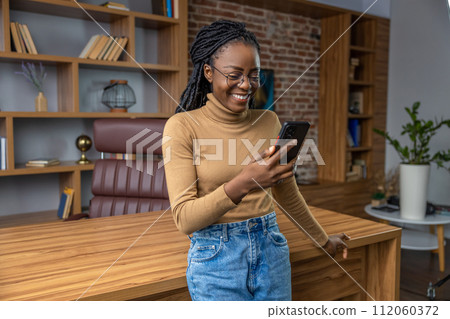 Cheerful woman using smartphone standing in office communicating online 112060372