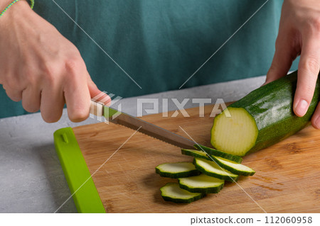 Woman's hands cutting zucchini on wooden cutting board at home, preparing healthy food, close up Woman's hands cutting zucchini on wooden cutting board at home, preparing healthy food, close up 112060958