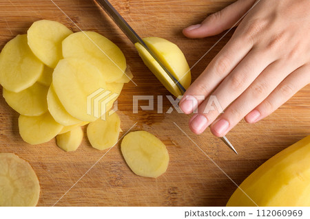 Authentic female hands cutting potatoes on wooden cutting board on gray kitchen table Authentic female hands cutting potatoes on wooden cutting board on gray kitchen table 112060969