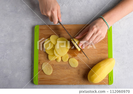 Authentic female hands cutting potatoes on wooden cutting board on gray kitchen table Authentic female hands cutting potatoes on wooden cutting board on gray kitchen table 112060970