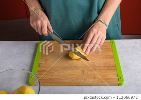 Authentic female hands cutting potatoes on wooden cutting board on gray kitchen table Authentic female hands cutting potatoes on wooden cutting board on gray kitchen table 112060978