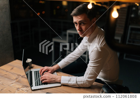 Portrait of serious focused young man working inside office with laptop at late night in dark room. Confident successful businessman thoughtful looking at camera. Concept overwork and deadline. 112061808