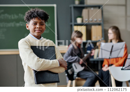 African American boy holding sheet music in folder standing in classroom, his classmate with bass guitar and teacher sitting in background 112062175