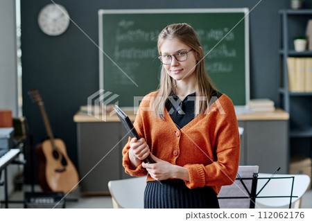 Medium portrait of young Caucasian woman working at music teacher holding folder with sheet music standing in classroom looking at camera Medium portrait of young Caucasian woman working at music teacher holding folder with sheet music standing in classroom looking at camera 112062176
