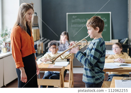 Selective focus shot of young female music teacher and kids listening to boy playing trumpet at class 112062255