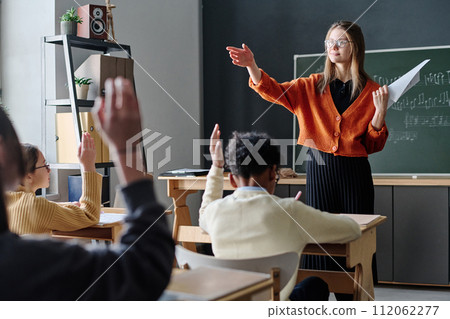 Young Caucasian female music teacher asking question, her students sitting at desks raising hands to answer it 112062277