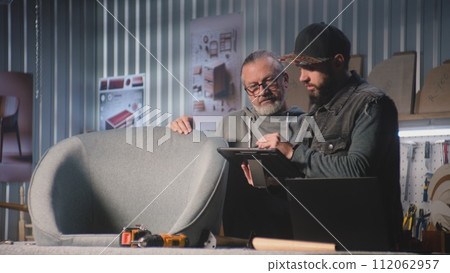 Carpenter or designer talks to colleague, uses tablet computer and laptop 112062957