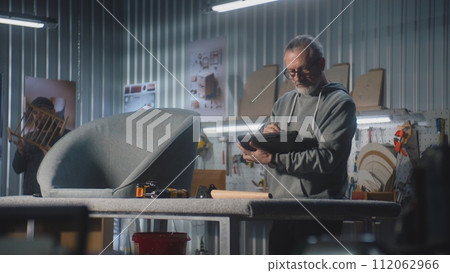 Mature carpenter or designer uses digital tablet in furniture workshop 112062966