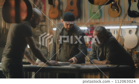 Tattooed craftsman holds guitar body and talks with colleagues 112063024