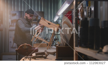 Male carpenter makes or assembles wooden bedside table 112063027