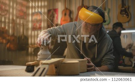 Mature craftsman in protective face shield polishes wooden guitar body Mature craftsman in protective face shield polishes wooden guitar body 112063061