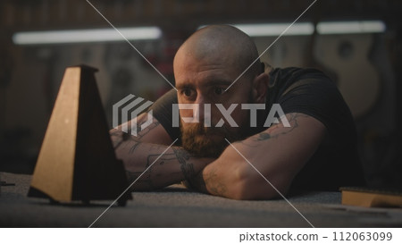 Middle aged tattooed craftsman sits at table in dark workroom Middle aged tattooed craftsman sits at table in dark workroom 112063099