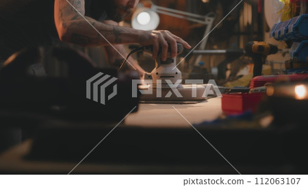 Male carpenter polishes wooden guitar body using polishing machine 112063107