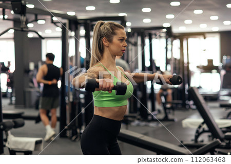 Muscular fitness woman athlete doing heavy weight exercise for shoulders with dumbbells in the gym Muscular fitness woman athlete doing heavy weight exercise for shoulders with dumbbells in the gym 112063468
