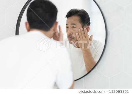 A middle-aged man in his 50s puts his hand on his cheek while looking in the mirror 112065501