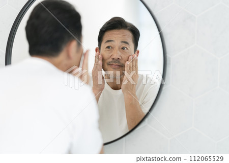 一名 50 多歲的男子照著鏡子，微笑著把手放在臉頰上。 112065529