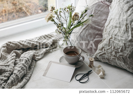 Easter composition. Spring still life. Cup of coffee, floral bouquet. Window sill. White tulips, carnation, wax flowers. Blank greeting card mockup. Pillows, old scissors. Scandinavian home, breakfast 112065672