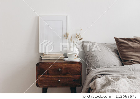 Vertical white picture frame mockup on old books. Elegant bedroom view. Brown, grey linen and velvet pillows, blanket. Wooden night stand with ceramic vase, dry grass and cup of coffee. Scandinavian Vertical white picture frame mockup on old books. Elegant bedroom view. Brown, grey linen and velvet pillows, blanket. Wooden night stand with ceramic vase, dry grass and cup of coffee. Scandinavian 112065673