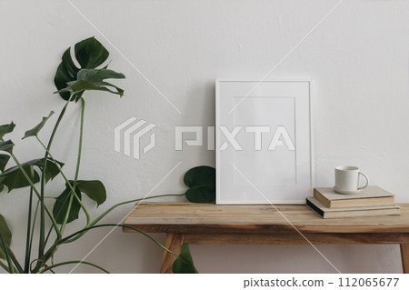 Vertical picture frame mockups on vintage wooden bench, table. Cup of coffee, tea on pile of books. Green monstera potted cheese plant. White wall background. Neutral Scandinavian interior, artistic 112065677
