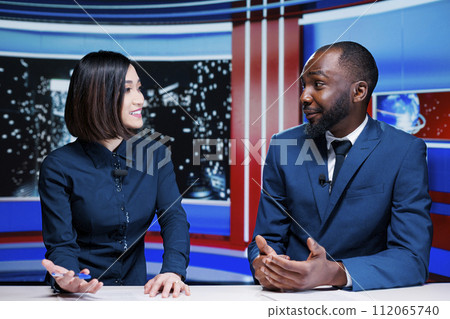 Two reporters broadcasting live information on international television channel, addressing all latest events and giving updates. Diverse presenters creating tv content in studio. 112065740