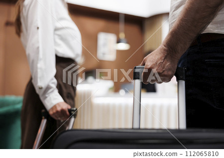 Close view of couple entering foyer of fancy hotel with trolley bags. Detailed shot of elderly individuals grasping suitcases as they walk up to registration desk to check in with the receptionists. 112065810