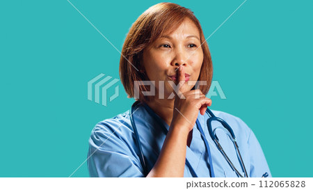 Close up shot of rigid strict BIPOC clinic manager shushing at camera, bothered by too much noise. Rigurous nurse holding quiet gesture finger to lips, isolated over studio background Close up shot of rigid strict BIPOC clinic manager shushing at camera, bothered by too much noise. Rigurous nurse holding quiet gesture finger to lips, isolated over studio background 112065828