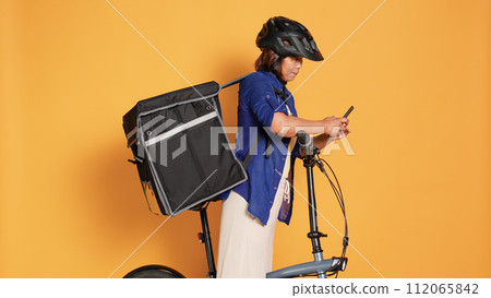 Asian cyclist delivering takeaway food to customers, imputing next location data in phone app. Courier woman riding bike, wearing safety helmet and thermal backpack, isolated over studio background 112065842