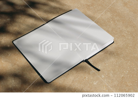 Tropical summer mockup. Open diary, book with black cover in sunlight. Blank pages. Harsh tree branches shadows. Beige marble stone texture, background. Holiday template. Flat lay, top view, no people 112065902