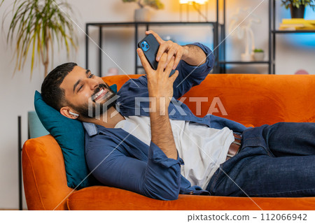 Indian man taking wireless earphones out of box listening to music enjoying leisure relaxing at home 112066942