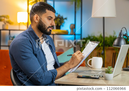 Indian man freelancer making notes on clipboard at home office using laptop prepare business report Indian man freelancer making notes on clipboard at home office using laptop prepare business report 112066966