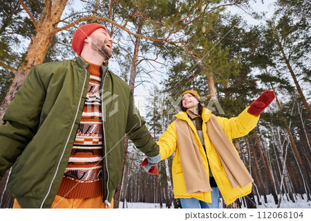 Low angle view of cheerful couple wearing stylish warm clothes holding hands enjoying walk in forest park on winter day 112068104