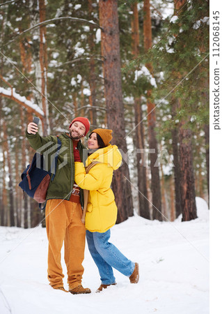 Vertical long shot of young Caucasian man and woman wearing down jackets taking selfie on smartphone in forest on cold winter day Vertical long shot of young Caucasian man and woman wearing down jackets taking selfie on smartphone in forest on cold winter day 112068145