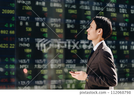 一名 30 多歲的男子在股價板前使用智慧型手機應用程式交易股票，並考慮使用 NISA。 112068798