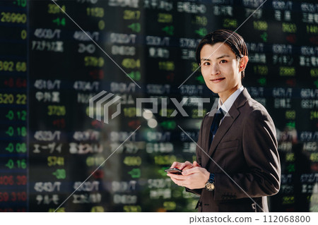 一位年輕的男性商人看著鏡頭,使用智慧型手機應用程式在 NISA 購買、出售和管理股票。 一位年輕的男性商人看著鏡頭,使用智慧型手機應用程式在 NISA 購買、出售和管理股票。 112068800