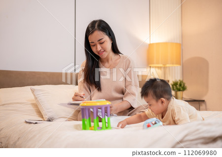 A cute Asian baby boy is playing with toys in bed while his mom is focusing reading documents. A cute Asian baby boy is playing with toys in bed while his mom is focusing reading documents. 112069980
