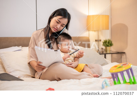 A busy Asian mom is holding her baby boy while talking on the phone and working on documents in bed. A busy Asian mom is holding her baby boy while talking on the phone and working on documents in bed. 112070037