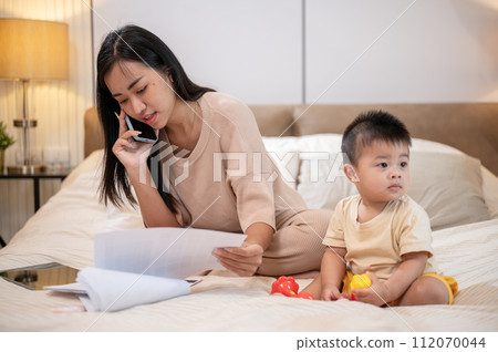 A cheerful, innocent Asian baby boy is playing toys in bed while his mom is talking on the phone. 112070044