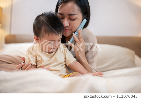 A cute, innocent Asian little boy is playing in bed while his mom is talking on the phone. 112070045