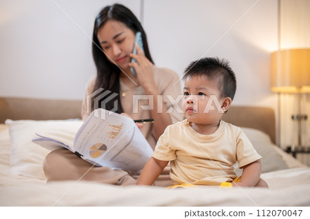A cute Asian little boy is playing toys in bed while his mother is working and talking on the phone. 112070047