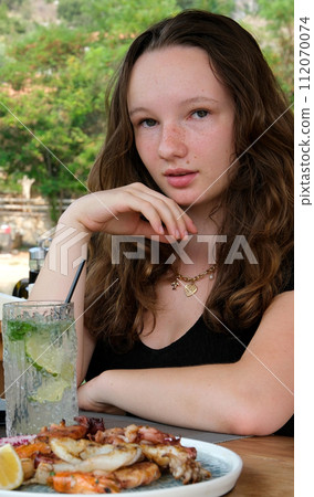 Young woman in a restaurant mojito and seafood on a plate against the backdrop of green river in Albania delicious food cooking relax straightening her hair black dress loose brown hair. High quality Young woman in a restaurant mojito and seafood on a plate against the backdrop of green river in Albania delicious food cooking relax straightening her hair black dress loose brown hair. High quality 112070074