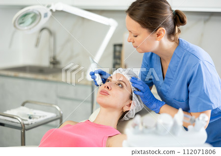 Adult woman cosmetologist doing microcurrent procedure to young woman 112071806