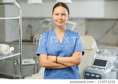 Portrait of female doctor cosmetologist in office of hardware cosmetic procedure. Beautiful beautician in uniform smiling, looking at camera and posing. In background Rf cosmetology device 112071818