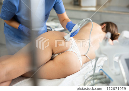 Adult woman cosmetologist gives ultrasonic massage of butt to young woman 112071959
