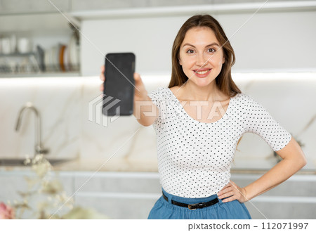 In kitchen, smiling girl in casual clothes shows blank smartphone screen close-up 112071997