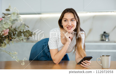 Middle-aged woman looking at her phone leaning on kitchen table 112072058