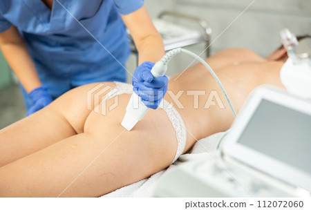 Lady lying on couch during treatment procedures with technologies vibrating shovel leather knife. Soften skin keratin, deep clear skin dirt and anti-cellulite massage buttocks. Hardware cosmetology 112072060