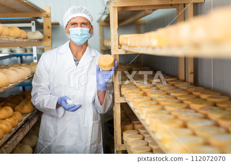 Cheesemaker in mask controlling maturing process of cheese wheels Cheesemaker in mask controlling maturing process of cheese wheels 112072070