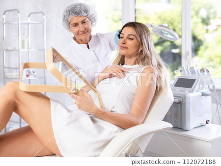 Satisfied young blonde woman looking in the mirror after plastic surgery. Consultation with professional surgeon Satisfied young blonde woman looking in the mirror after plastic surgery. Consultation with professional surgeon 112072188